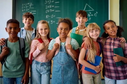 Seven diverse children standing together in classroom with chalkboard behind them, smiling at camera.