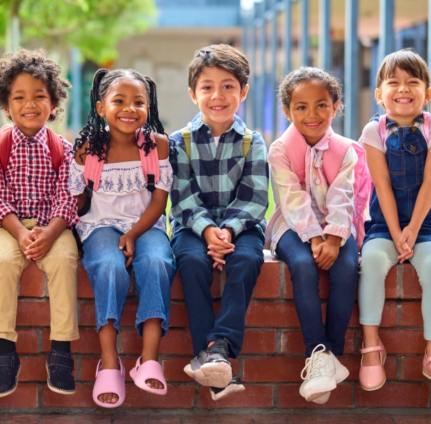 Five diverse children sitting together on brick steps outside a school building, smiling at camera.