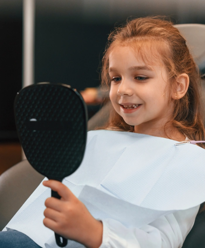 Young girl with reddish hair smiling while holding a black paddle mirror in dental office.