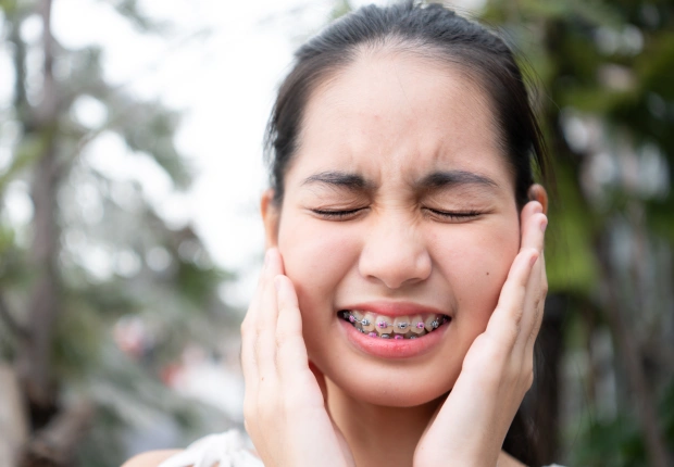 Young girl with braces experiencing tooth pain, holding cheeks outdoors near trees.