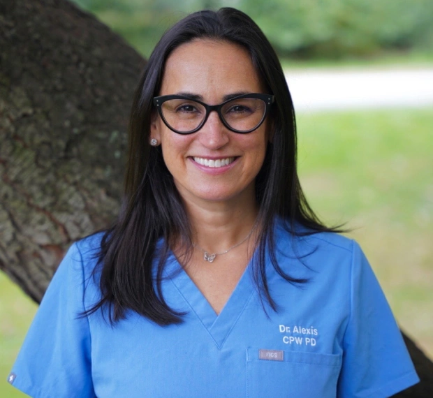 Female dental professional in blue scrubs smiling outdoors against tree, wearing glasses.