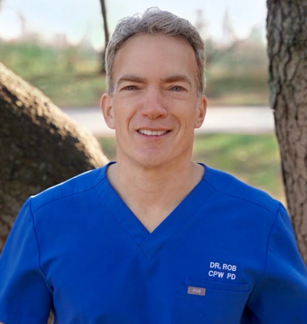 Dr. Rob in blue scrubs smiling outdoors by trees near water.