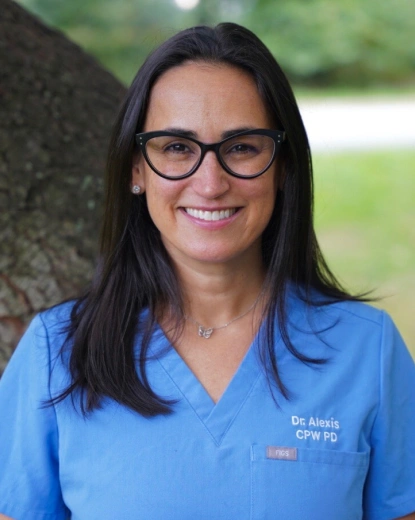 Dr. Alexis smiling outdoors in blue scrubs against a tree background.