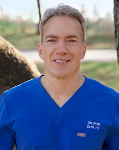 Dr. Rob smiling outdoors wearing blue scrubs with waterfront background