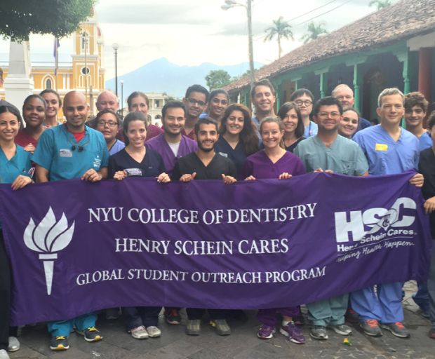 NYU dental students holding Henry Schein Cares banner during global outreach program in tropical setting.