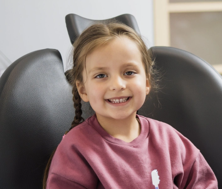 Young girl smiling in dental chair wearing pink shirt at pediatric dental office.