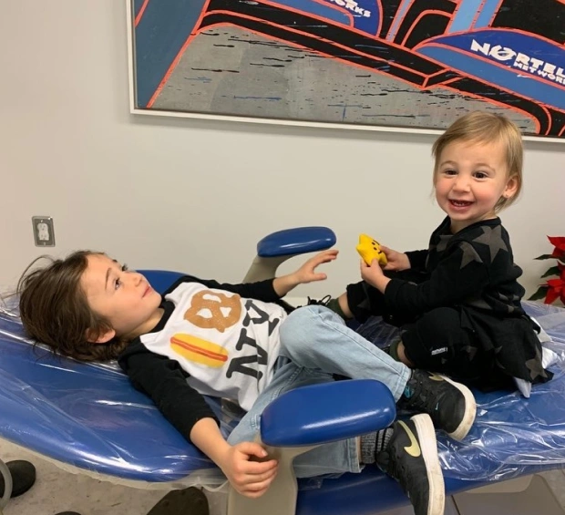 Two young children playing together on a blue dental chair in a pediatric dental office.