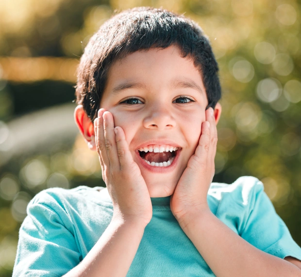 Young boy smiling brightly with hands on cheeks outdoors in natural sunlight.