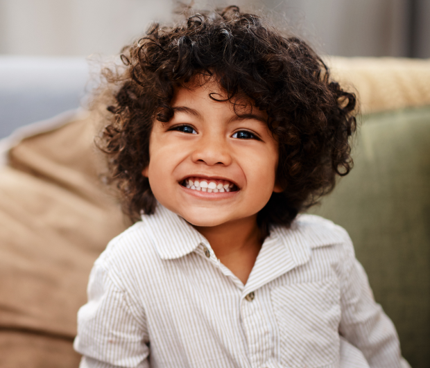 Young child with curly dark hair smiling brightly in home setting on couch.