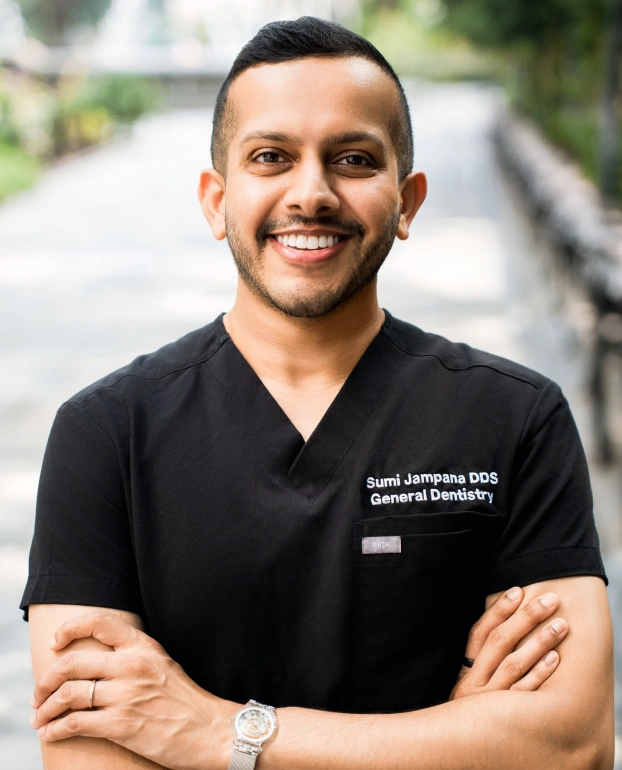 Portrait of Dr. Sumi Jampana, DDS, smiling with arms crossed, wearing black scrubs with "Sumi Jampana DDS General Dentistry" embroidered on the pocket.