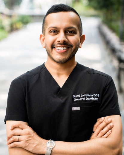 Portrait of Dr. Sumi Jampana, a general dentist, smiling outdoors with arms crossed, wearing black medical scrubs labeled "Sumi Jampana DDS General Dentistry".